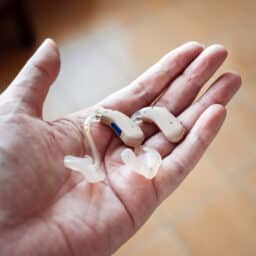 A hand holding a pair of modern hearing aids against the background of a tile floor