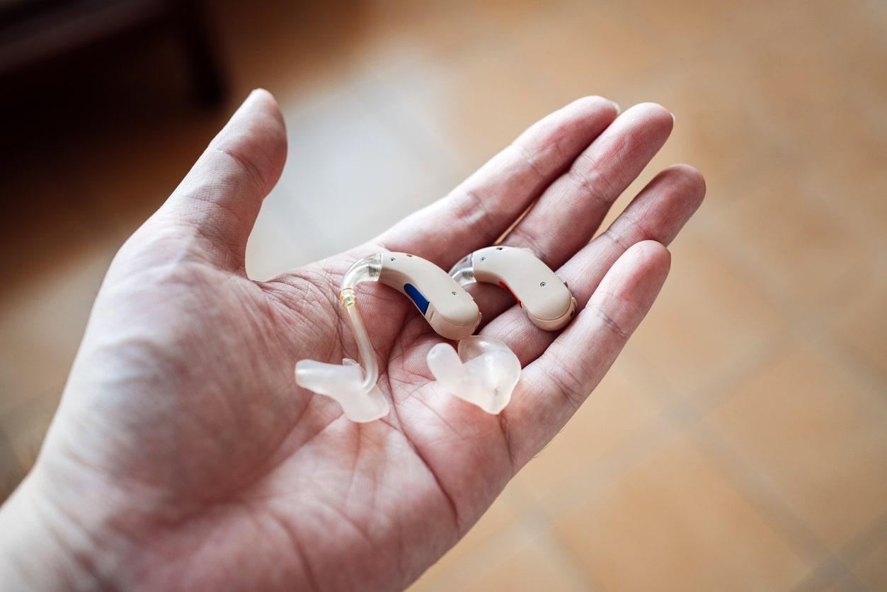A hand holding a pair of modern hearing aids against the background of a tile floor