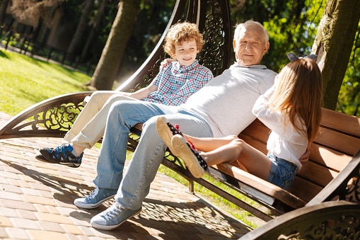 Grandfather with his two grandkids sitting on a bench in a park on a beautiful day.