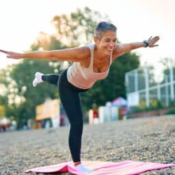 Smiling mature woman practicing yoga warrior three pose on a pink mat in a park, enjoying a healthy lifestyle and promoting physical and mental wellness