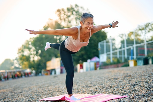 Smiling mature woman practicing yoga warrior three pose on a pink mat in a park, enjoying a healthy lifestyle and promoting physical and mental wellness