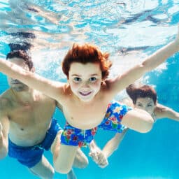 Happy child swimming in a blue pool with their family. Children with hearing loss need to remove their hearing aids before swimming to avoid damage.