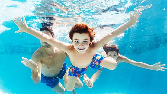 Happy child swimming in a blue pool with their family. Children with hearing loss need to remove their hearing aids before swimming to avoid damage.